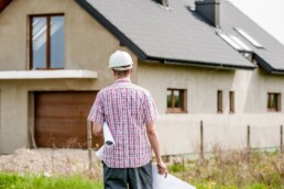 A man wearing a hard hat and holding plans walks towards a custom home.