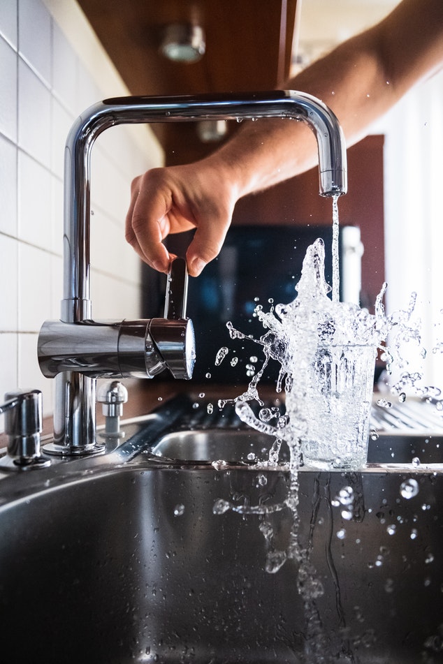 A hand runs a modern faucet, causing a glass of water to overflow.